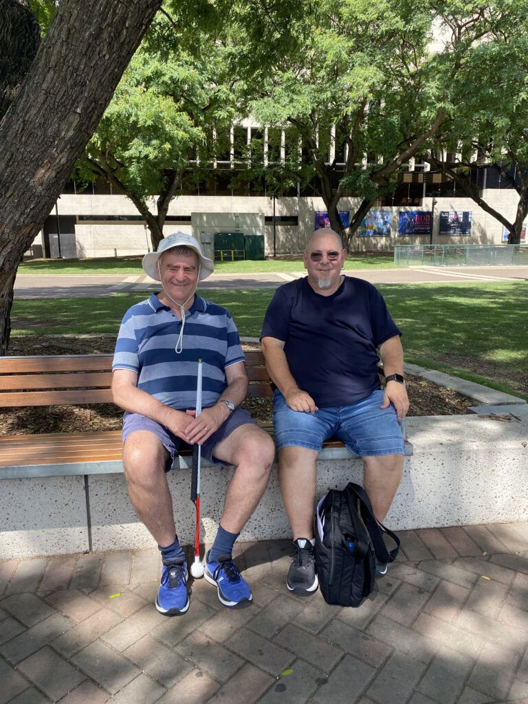 Joe and one of his support workers, Shawn, sitting on a park bench under a big tree on a sunny day.