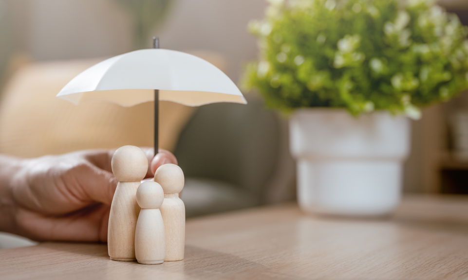 Hand sheltering small wooden figures of a family under a white umbrella, symbolising family protection and care