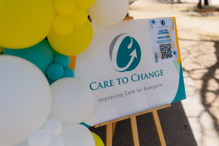 Care to Change 'Improving Care for Everyone' sign on an easel framed by balloons at a community event