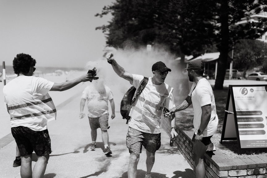 Black and white photo of participants tossing colour powder beside event signage at a beachside Walk for Awareness event